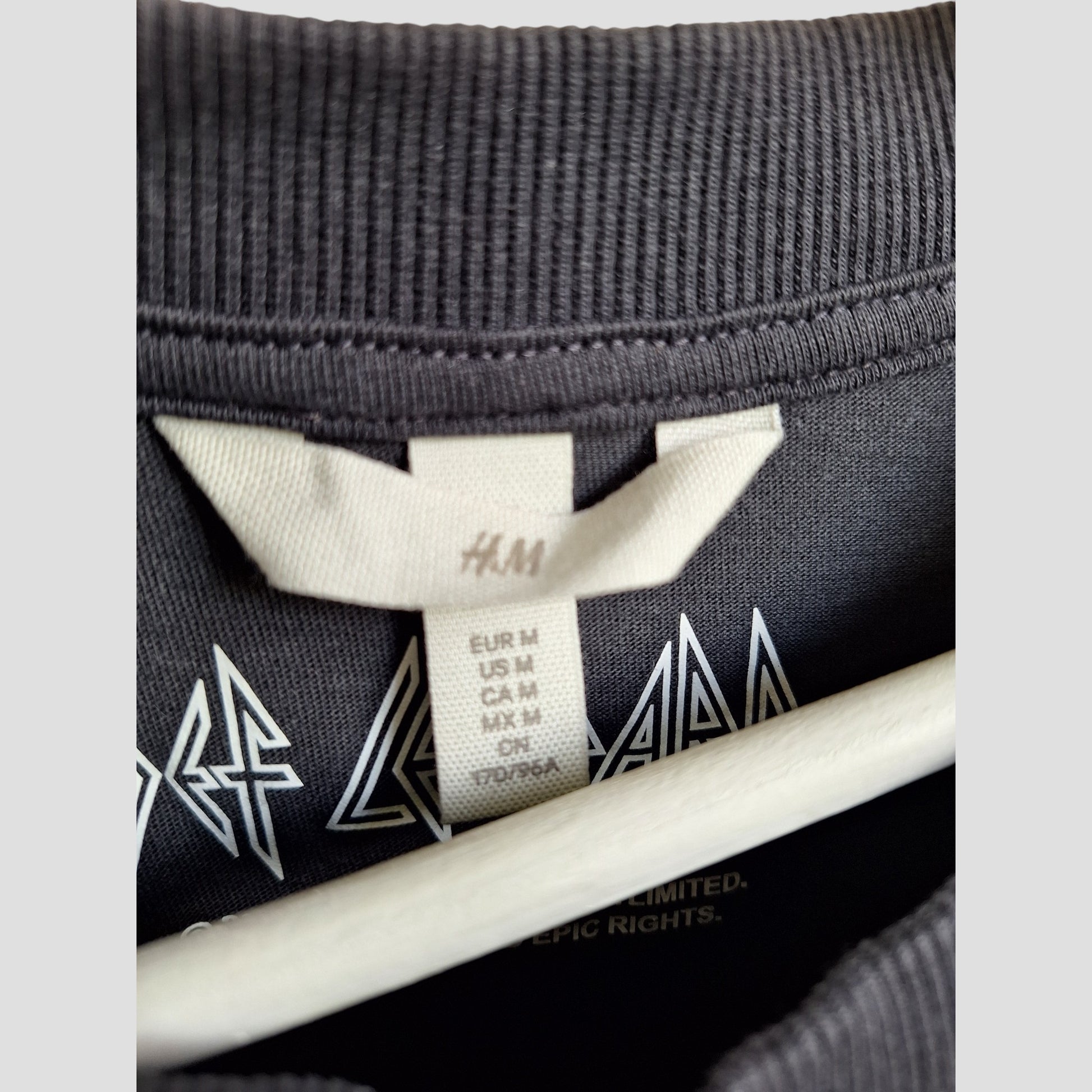 Close-up of an H&M tag sewn into the Upcycled Breastfeeding & Pumping Grey Def Leppard Band t-shirt M (100370), showing size markers (EUR/US/CA/MX M, CN 170/96A) with the shirt’s graphic partly visible.
