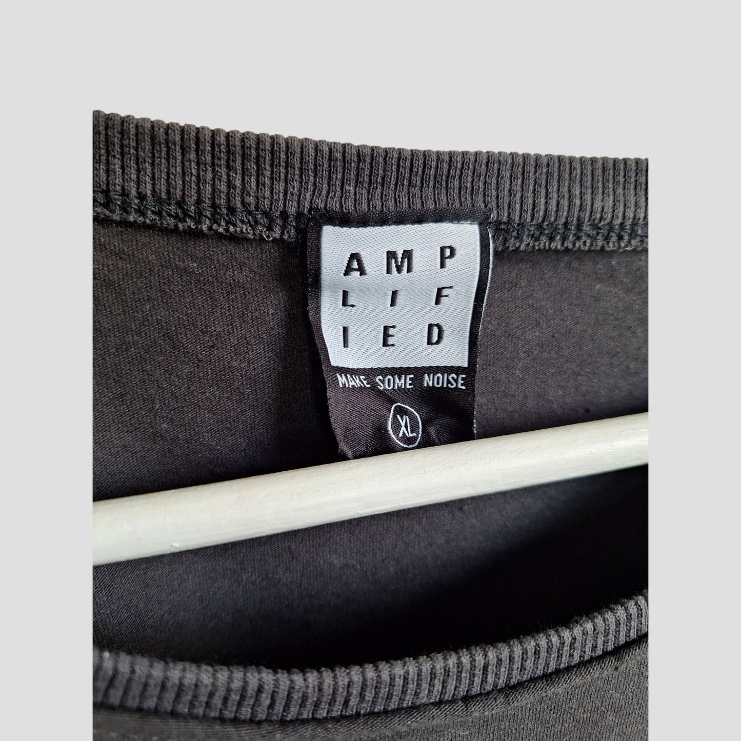 A close-up of a black label inside an upcycled XL Amplified Metallica Guitar Band nursing & pumping t-shirt shows the brand, MAKE SOME NOISE phrase, and size XL. Part of a hanger is visible beneath the label.