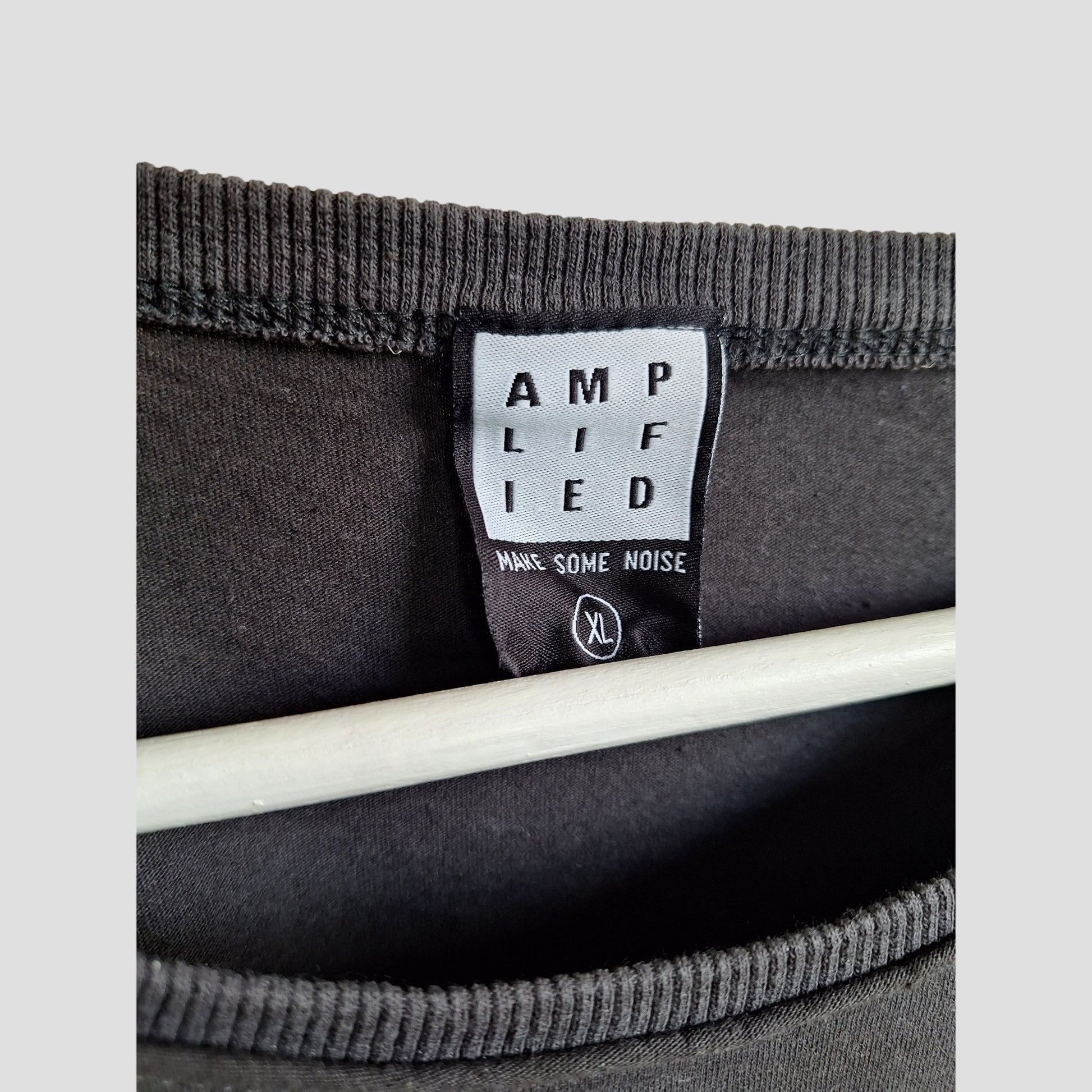 A close-up of a black label inside an upcycled XL Amplified Metallica Guitar Band nursing & pumping t-shirt shows the brand, MAKE SOME NOISE phrase, and size XL. Part of a hanger is visible beneath the label.