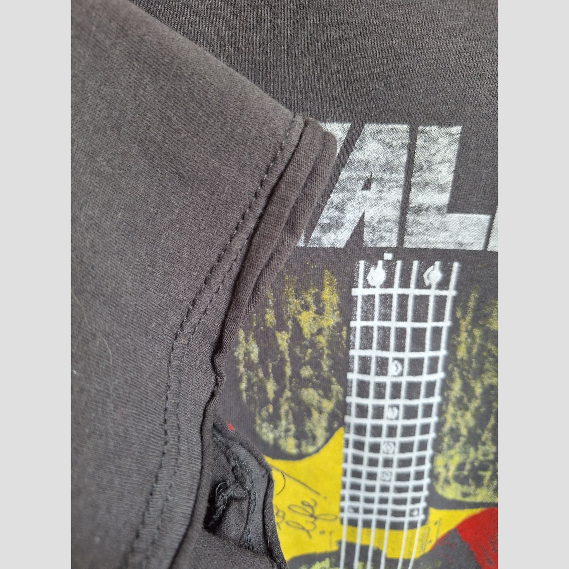 Close-up of an upcycled Amplified XL Metallica Guitar Band breastfeeding & pumping T-shirt, featuring a visible seam, neck and headstock graphic, partial white text with yellow, red, and green accents. Product code: 100353.