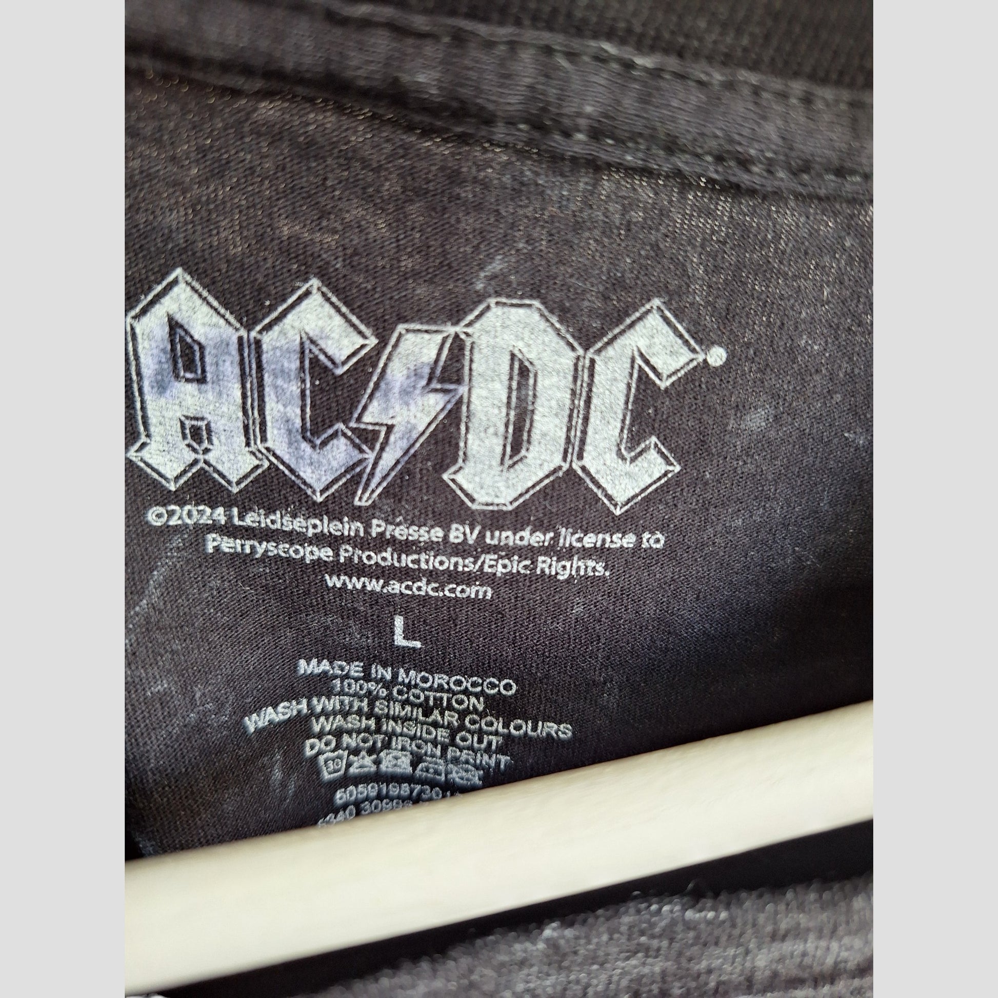 Close-up of the label on the Upcycled Breastfeeding & Pumping Black Floral AC/DC Band t-shirt (size L), showing the AC/DC logo, copyright info, website, care instructions, eco clothing details, and a wooden hanger at the bottom.