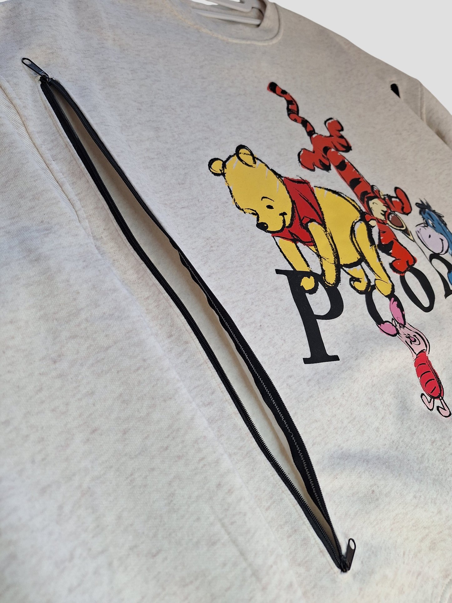 A close-up of a Primark Upcycled Nursing & Pumping Cream Winnie the Pooh Sweatshirt (XL, UK18/20) with a large diagonal zipper and “Pooh” text—an adorable preloved piece for breastfeeding moms.