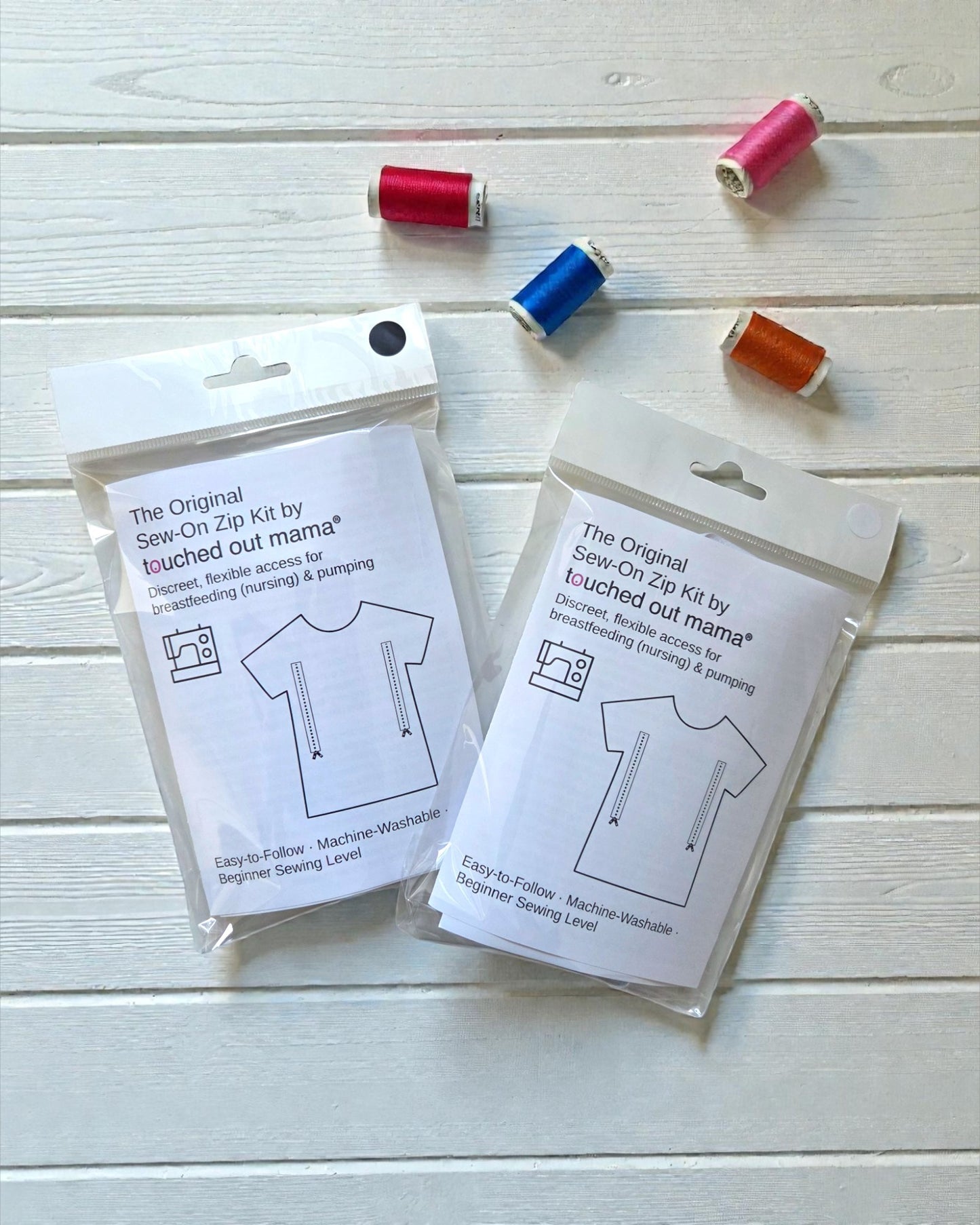 Two touched out mama Original Sew-On Zip Kits for Breastfeeding & Pumping Access are shown on a white wooden surface with five colorful spools of thread above them—a clever DIY nursing hack for moms.