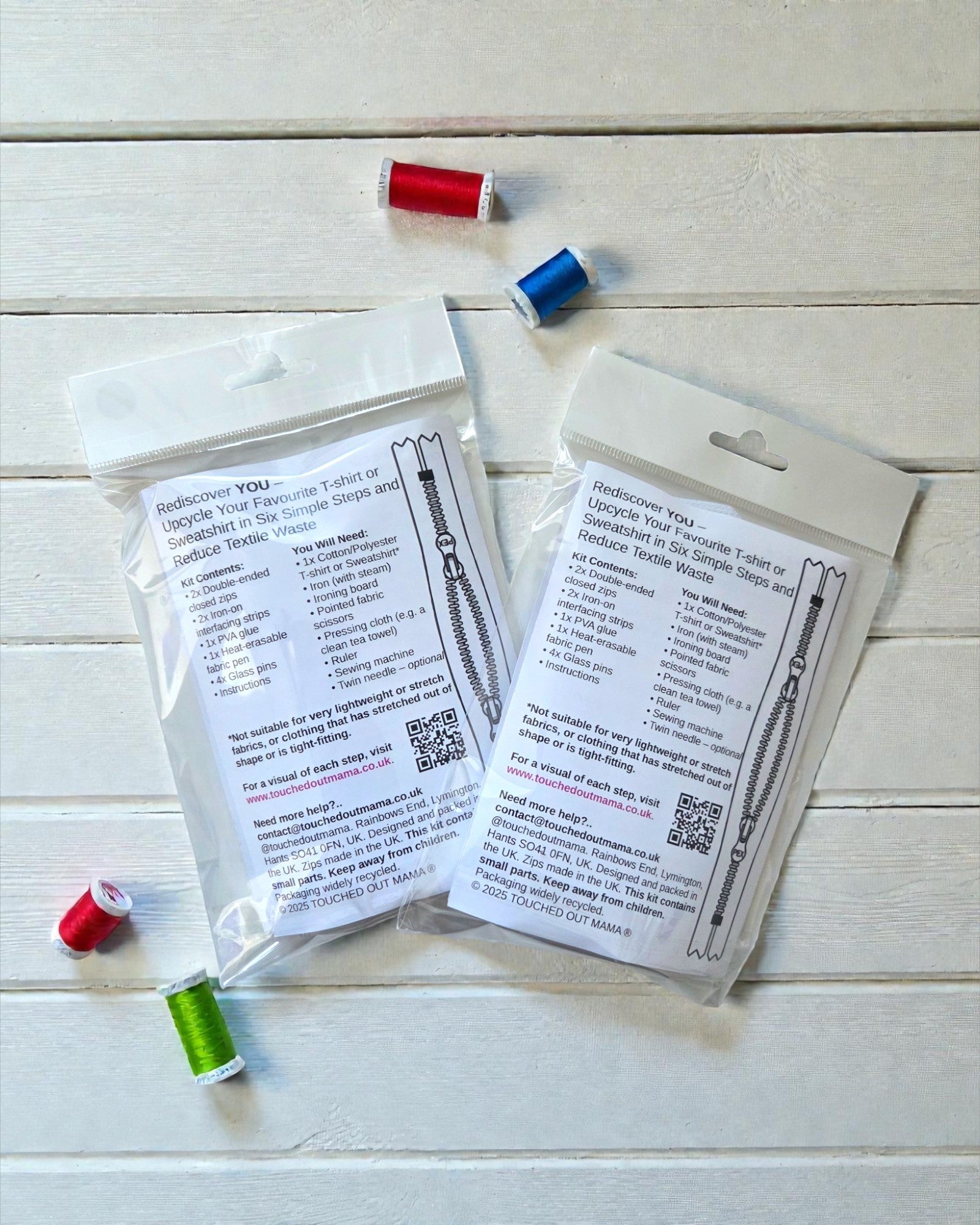 Two touched out mama Original Sew-On Zip Kits for Breastfeeding & Pumping Access are displayed on a white wooden surface with four small spools of red, blue, and green thread. Printed instructions are visible through the clear packaging.