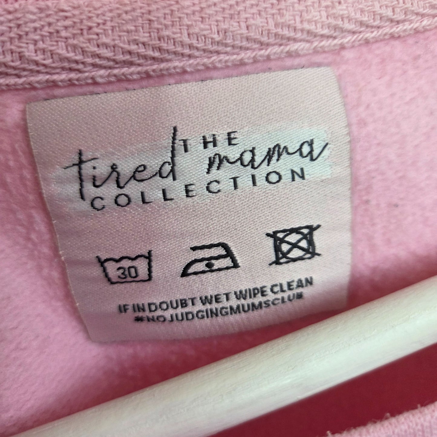 A clothing label on an upcycled pink breastfeeding & pumping sweatshirt (UK14, The Tired Mama Collection) features care symbols and the reminder: “If in doubt wet wipe clean #NOJUDGINGMUMSCLUB.”.