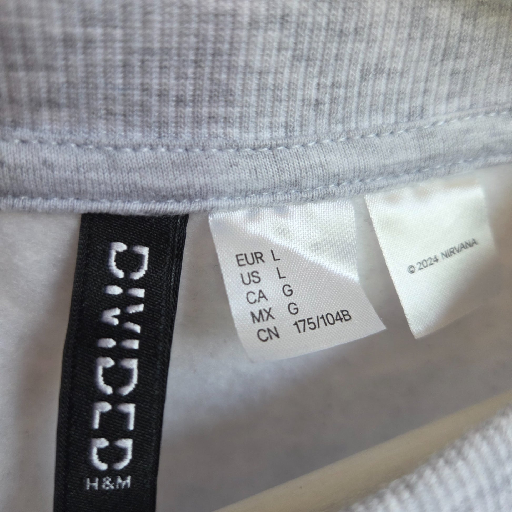 Close-up of labels on the H&M Upcycled Breastfeeding & Pumping Nirvana Band Sweatshirt (L, light grey/blue), showing international sizes and © 2024 Nirvana—emphasizing its eco-friendly design.