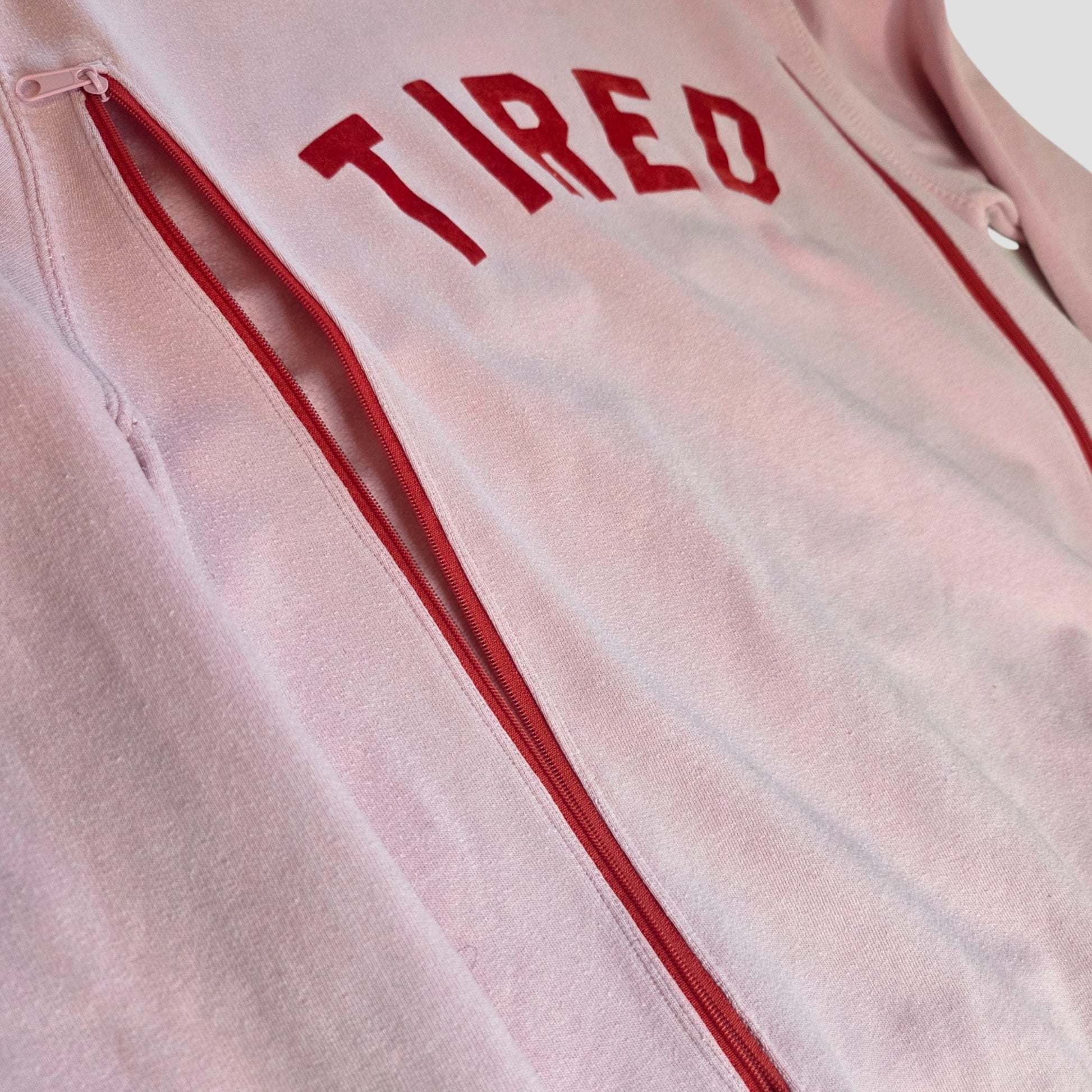 The Tired Mama Collection’s Upcycled Breastfeeding & Pumping Pink Tired Mama Sweatshirt UK14 (100429) features light pink fabric, bold red "TIRED" lettering, and red zippers—a standout piece for nursing and pumping moms.