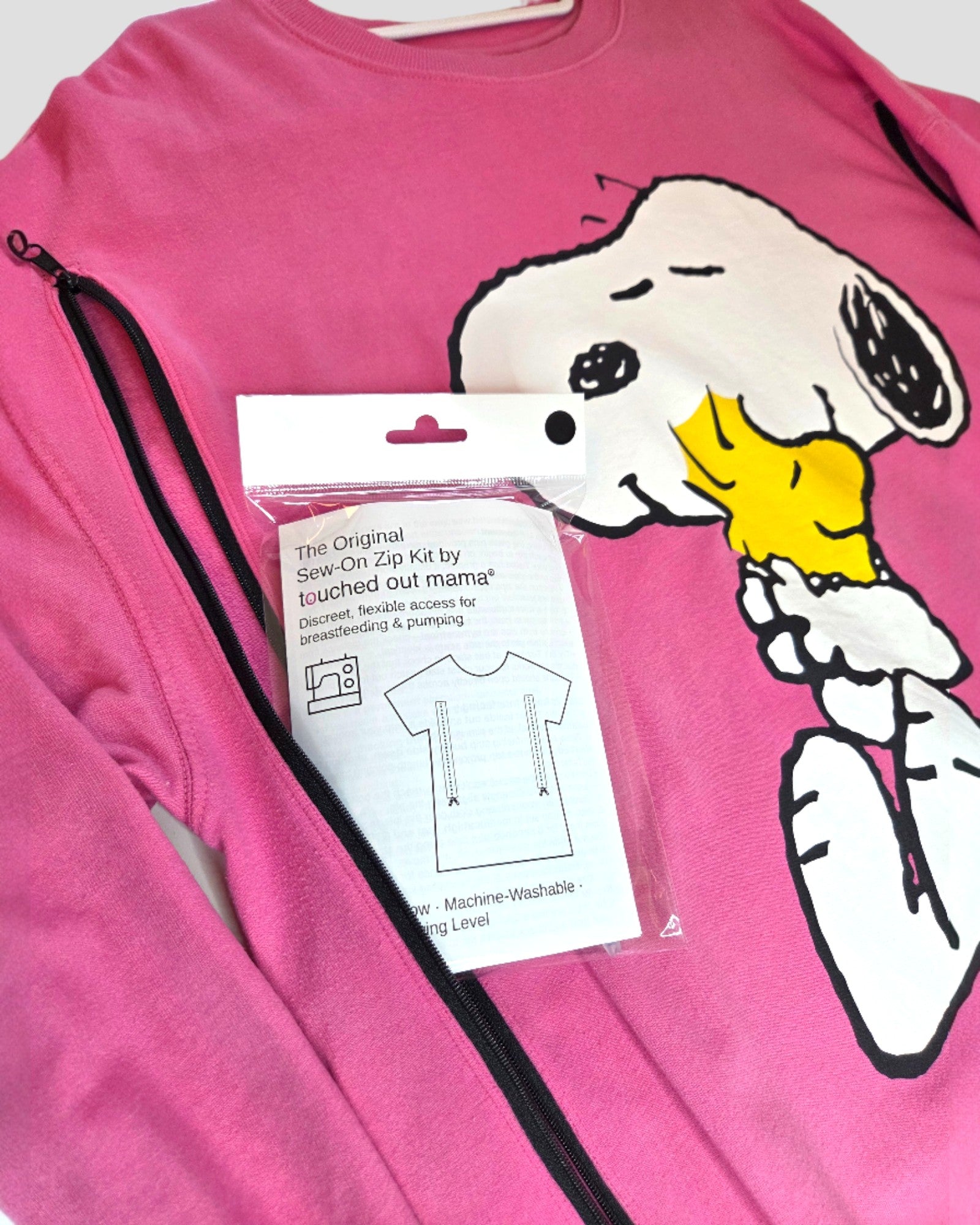 A pink sweatshirt with a Snoopy and Woodstock graphic displays a black zipper. On top is the "touched out mama The Original Sew-On Zip Kit for Breastfeeding (Nursing) & Pumping Access," perfect for upcycling or DIY nursing projects.