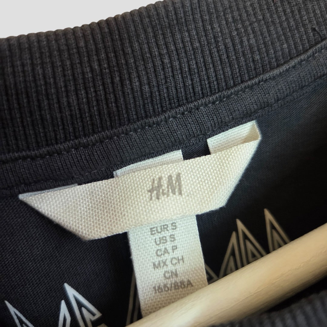Close-up of an H&M clothing label on a black garment, featuring size details and the collar with part of a wooden hanger. Showcases the Upcycled Breastfeeding & Pumping Grey Def Leppard Leopard Band t-shirt (S, 100474).
