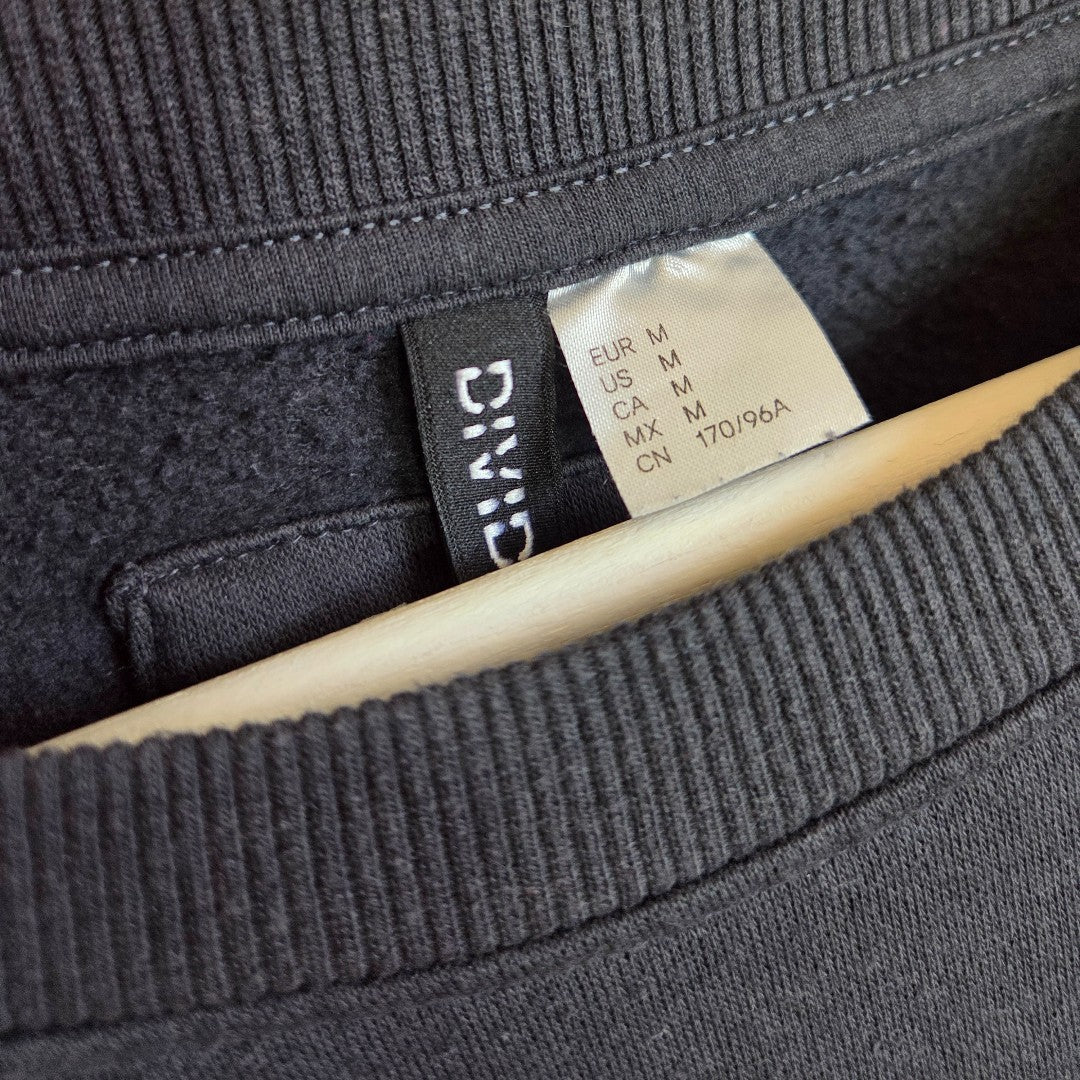 Close-up of a black clothing label on the H&M Upcycled Breastfeeding & Pumping The Doors Purple Band Sweatshirt (M, 100443), showing EUR/US/CA/CN size "M" and code "7701964," draped over a wooden hanger.