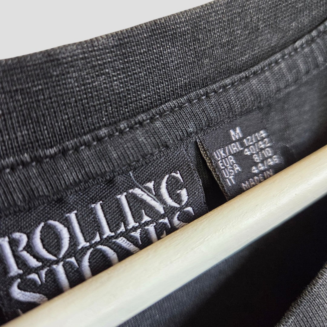 Close-up of a black Primark Upcycled Nursing & Pumping Rolling Stones Band t-shirt dress (M, UK12/14), hanging on a hanger. The neckline displays two tags: one with "ROLLING STONES," the other with size medium and care details.