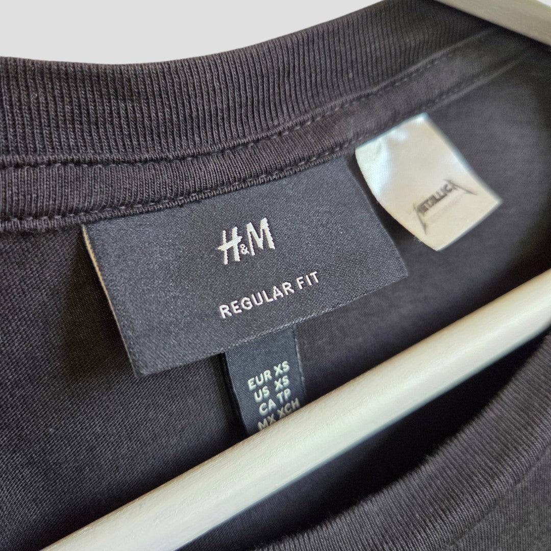 Close-up of an upcycled H&M black Metallica Skull Band maternity t-shirt (XS), with tags for the brand, Regular Fit, and sizing. Affordable, eco-friendly nursing/pumping top displayed on a white hanger.