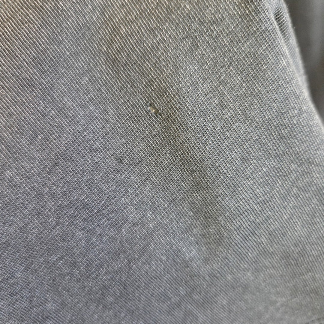 Close-up of a gray fabric from a Primark Upcycled Breastfeeding & Pumping The Doors Band t-shirt (M, UK12/14, 100326), showing fine woven texture, slight folds, a small hole, and minor thread pulls in the center.