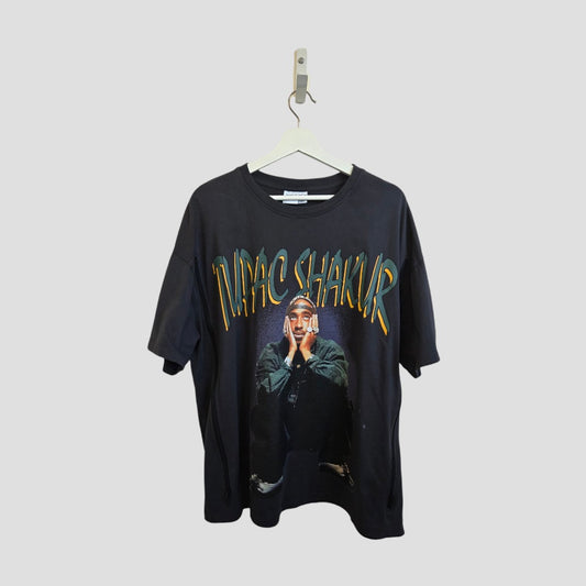 A grey upcycled Primark breastfeeding & pumping Tupac band t-shirt (L, UK14/16) hangs on a hanger, featuring a graphic of Tupac and bold yellow and green lettering above the image.