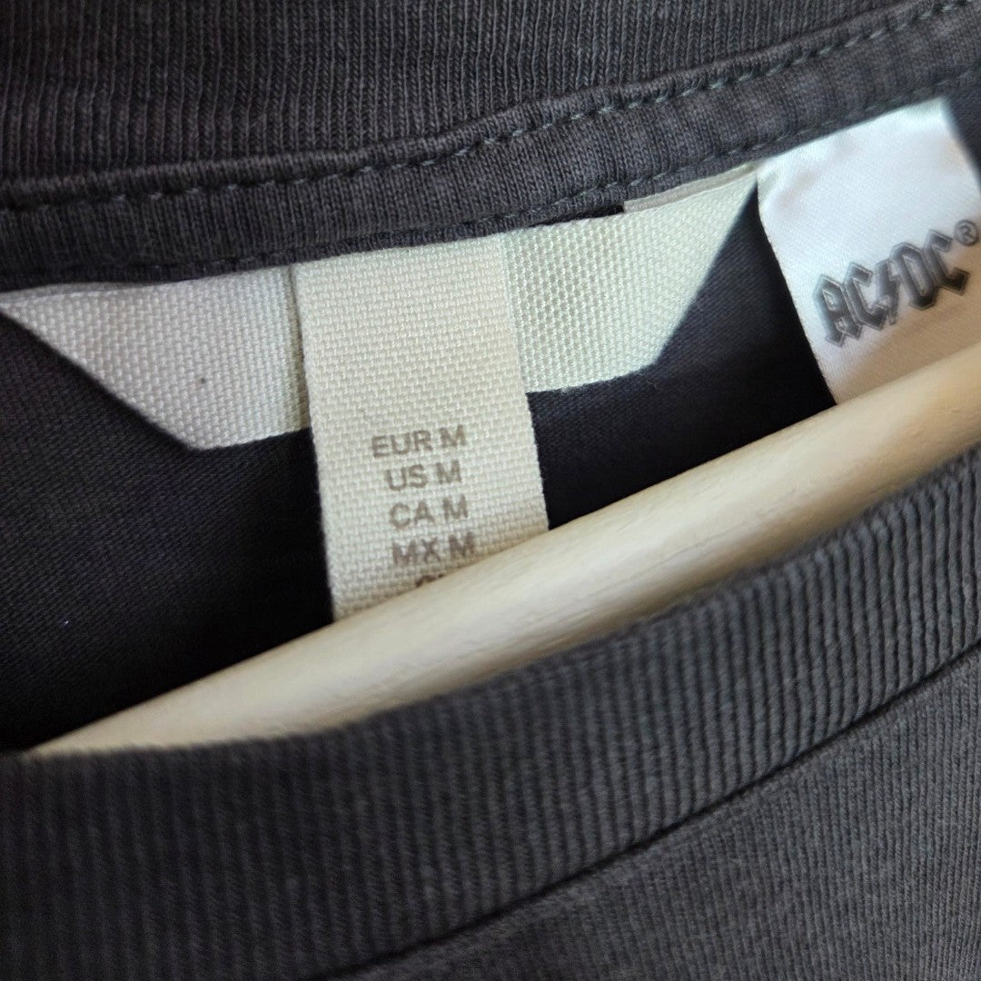 Close-up of the neck area of an upcycled H&M breastfeeding & pumping AC/DC Back in Black band t-shirt (M), dark grey, on a hanger. Two fabric tags: one with sizes and one partially showing the AC/DC logo on this eco-friendly piece.