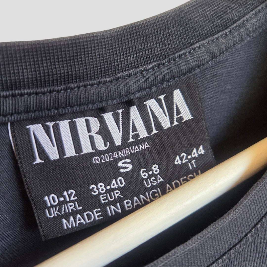 Close-up of a black Primark label inside the Upcycled Breastfeeding & Pumping Nirvana Turquoise Angel Band t-shirt (M, UK12/14), showing NIRVANA, size info, and Made in Bangladesh text. Wooden hanger visible underneath the label.