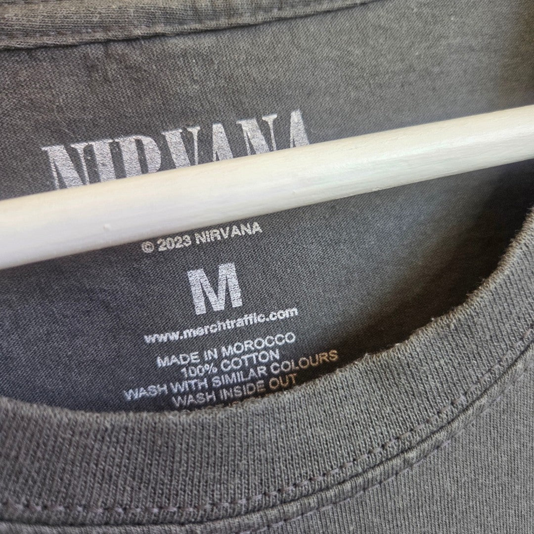 Close-up of a grey Nirvana Upcycled Breastfeeding & Pumping Guitar Band T-shirt (size M, 100% cotton), made in Morocco. Eco-friendly with care instructions and www.merchtraffic.com on the label; displayed on a hanger.