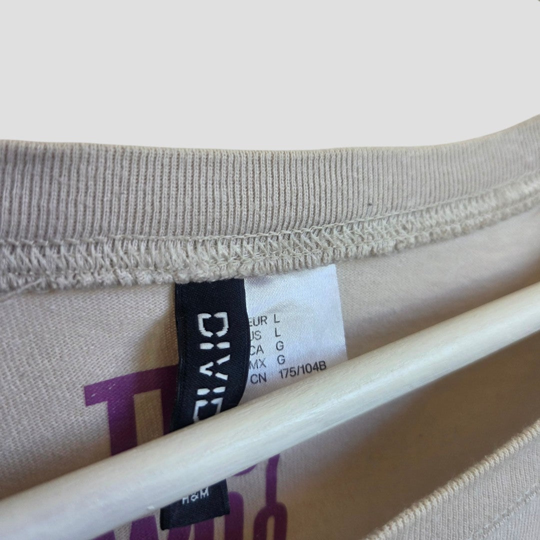 Close-up of an H&M Upcycled Breastfeeding & Pumping Cream/Pink The Who Band t-shirt (L) on a hanger shows the size tag with international codes and a hint of purple graphic, adding style to this affordable maternity essential.