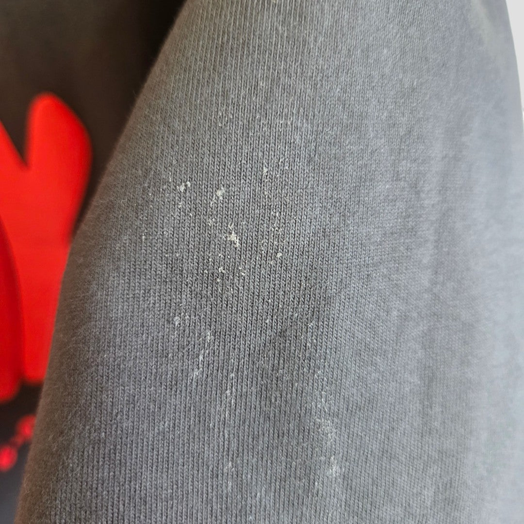 Close-up of a black fabric from the White Fox Upcycled Breastfeeding & Pumping Grey/Red t-shirt (S/M, 100495), showing small lint particles on the surface; a blurry red object appears in the background.