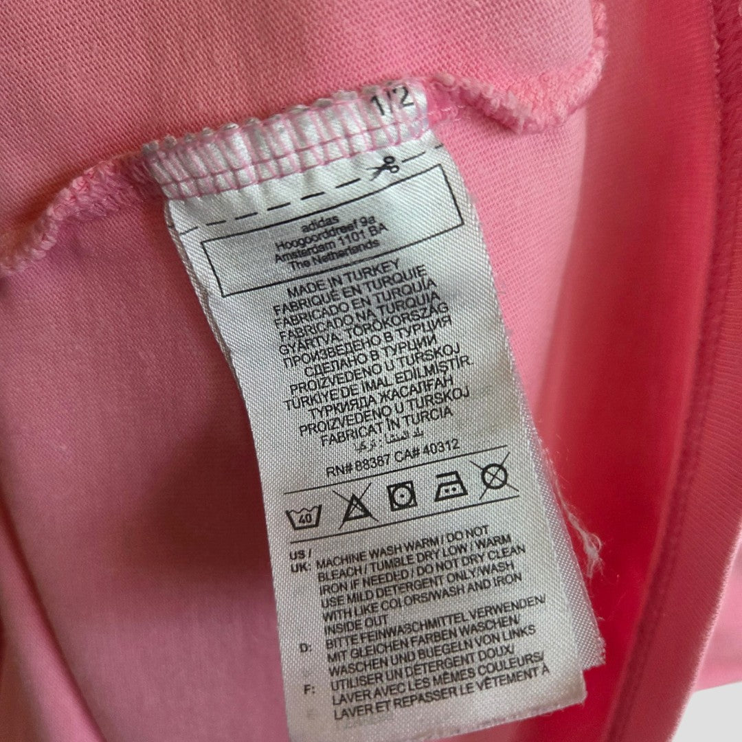 A close-up of a white care label on a pink Adidas Upcycled Breastfeeding & Pumping Retro t-shirt (L), showing multi-language instructions, brand info, and material details for this eco-friendly garment.