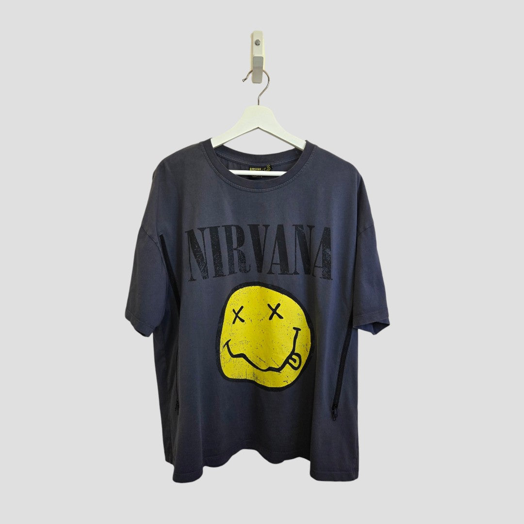 An upcycled Primark Nirvana band T-shirt (size L, UK14/16) hangs on a white hanger. It's grey with "NIRVANA" in large letters above a yellow, distressed smiley face with X eyes. The plain background is light gray.