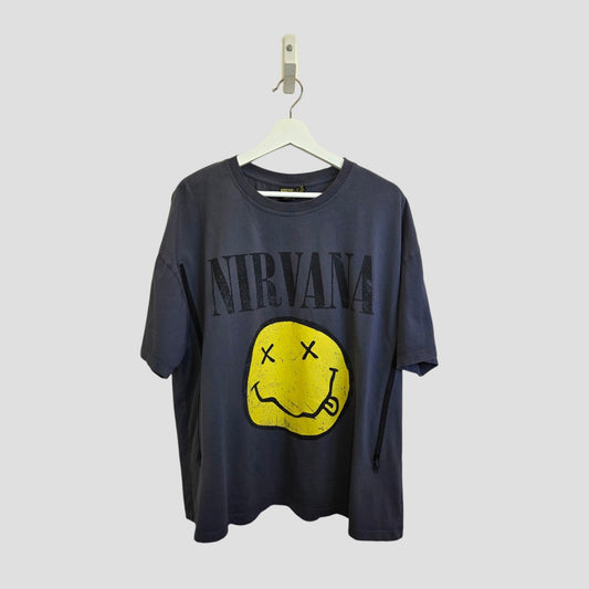 An upcycled Primark Nirvana band T-shirt (size L, UK14/16) hangs on a white hanger. It's grey with "NIRVANA" in large letters above a yellow, distressed smiley face with X eyes. The plain background is light gray.