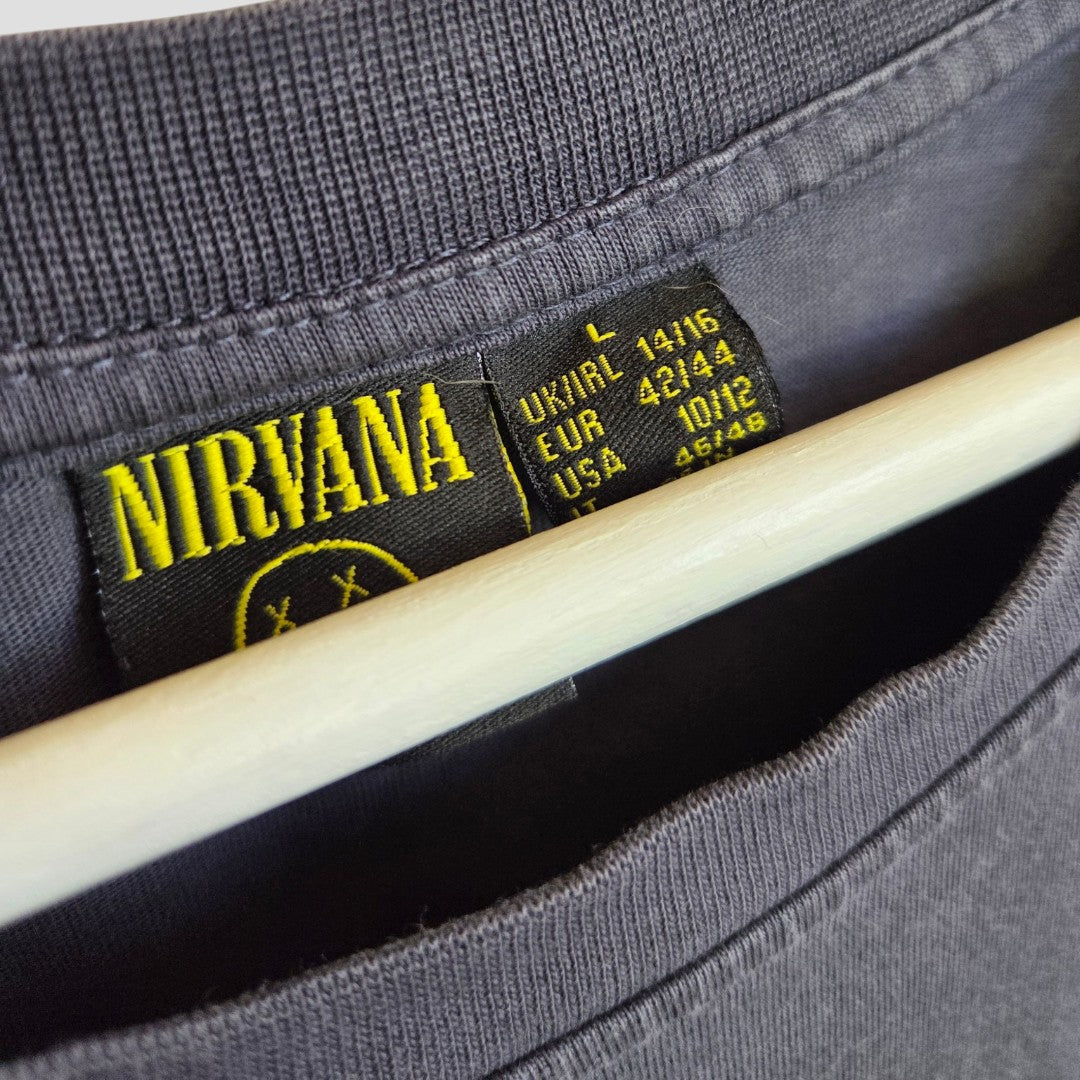 Close-up of an affordable Primark Upcycled Breastfeeding & Pumping Nirvana Band T-shirt in grey with a yellow smiley face, size L (UK14/16), hanging on a wooden hanger, featuring UK, EUR, and USA size details on the tag.