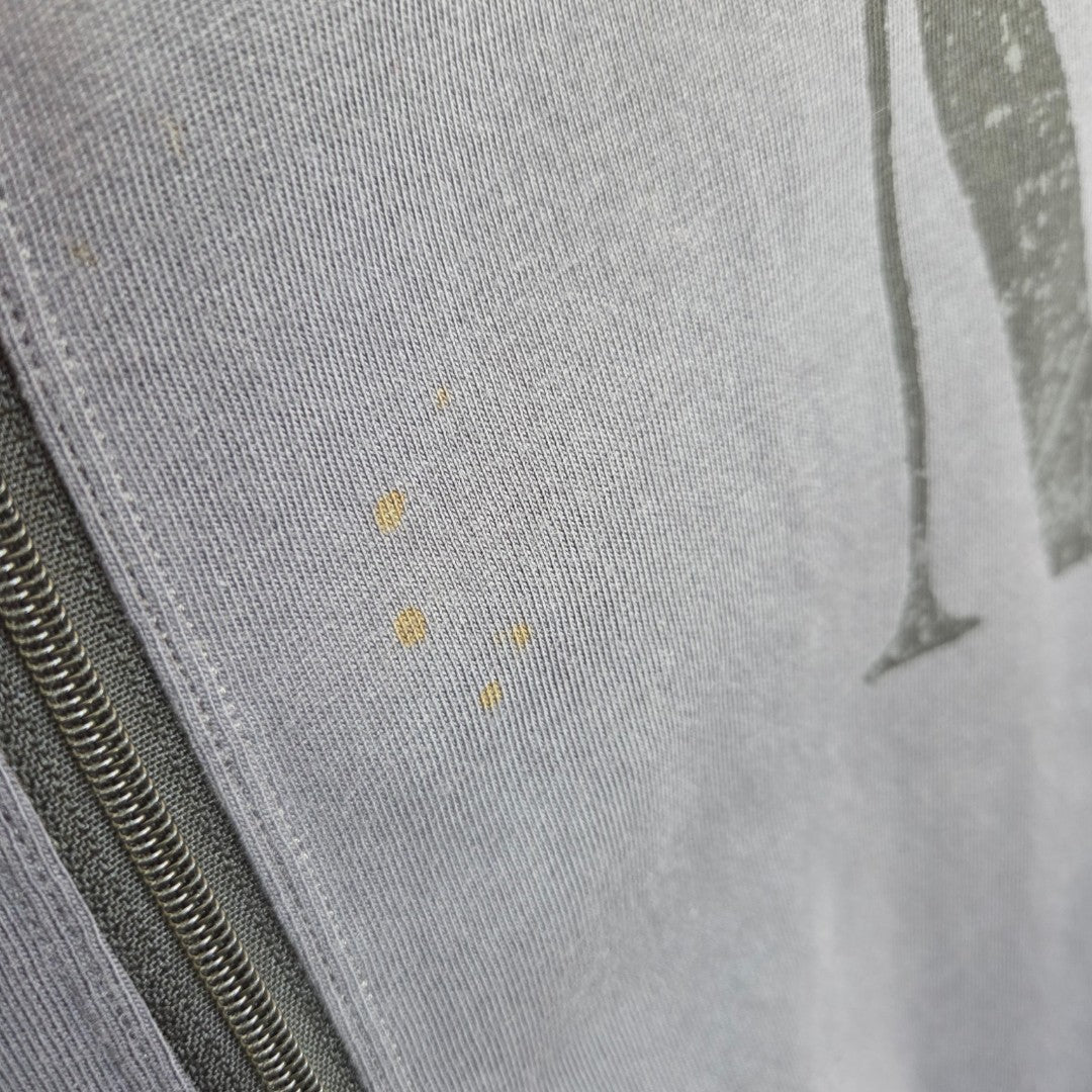 A close-up of the affordable Primark Upcycled Breastfeeding & Pumping Grey Yellow Smile Nirvana Band t-shirt (L, UK14/16) shows gray fabric with a left zipper, faint yellow stains near the center, and a partial black design on the right.