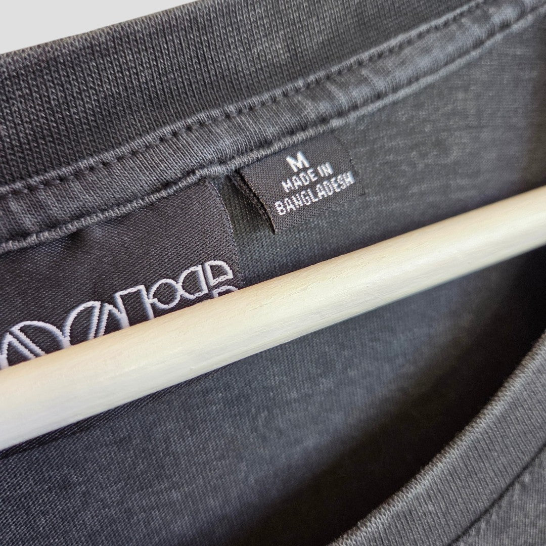 Close-up of an Upcycled Breastfeeding & Pumping The Doors Strange Days Band t-shirt (M, Primark, 100436) in black on a hanger, showing a clothing label with white text reading "M Made in Bangladesh.