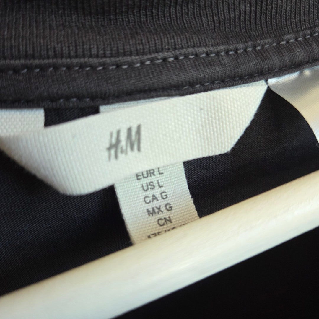 A close-up of an H&M label on the Upcycled Breastfeeding & Pumping Grey/Pink Nirvana Angel Band t-shirt (L), showing sizes EUR L, US L, CA G, MX G, and CN on a dark garment with the tag hanging from a white hanger.