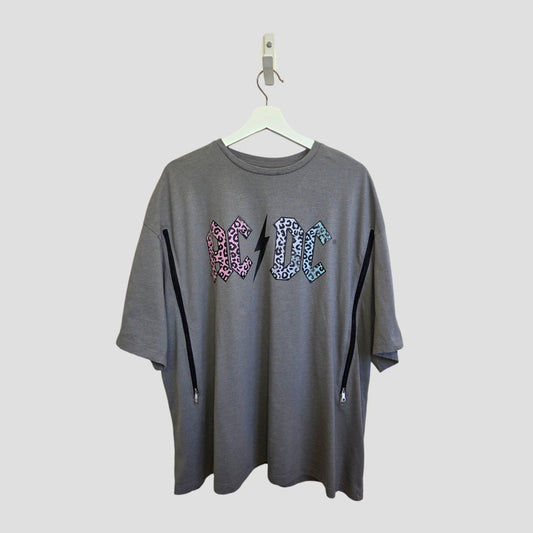 A loose-fitting Upcycled Breastfeeding & Pumping Grey Leopard AC/DC Band t-shirt by Tu (UK22) hangs on a white hanger, featuring diagonal zippers for easy nursing and colorful patterned letters on the chest.