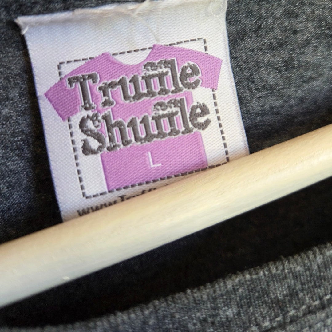 A label with a purple shirt icon and "Truffle Shuffle L" is sewn onto dark gray fabric from the Upcycled Breastfeeding & Pumping The BFG t-shirt (L) by Truffle Shuffle. A wooden hanger peeks out below the label.