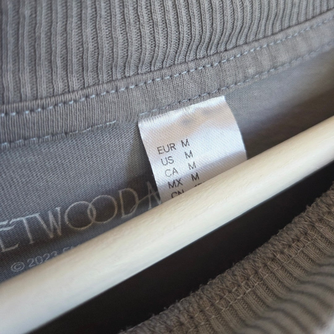 A close-up of an upcycled H&M Breastfeeding & Pumping Grey/Blue Fleetwood Mac Dreams Band t-shirt (size M, 100487), hanging on a white wooden hanger.