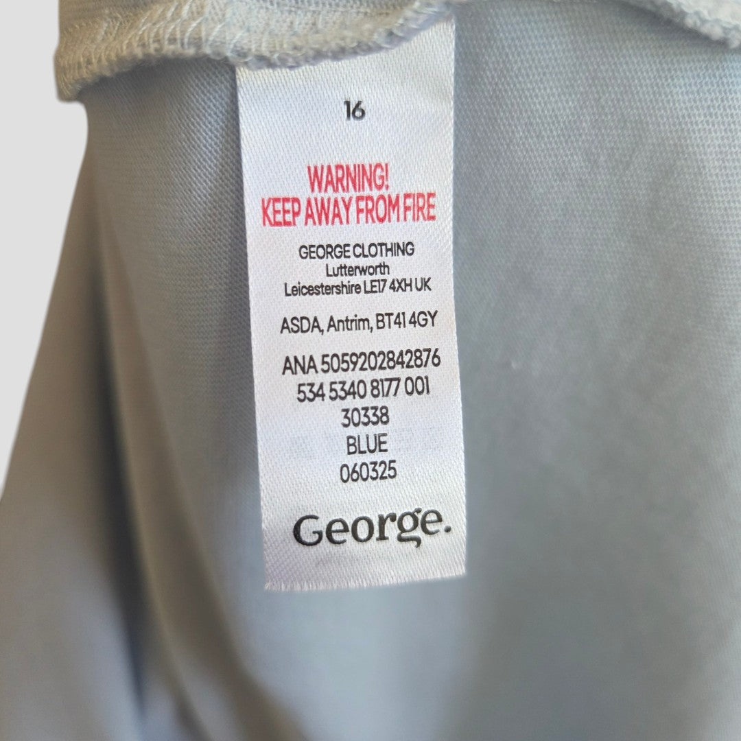 A close-up of a label with red text "KEEP AWAY FROM FIRE" and black text showing product info: George, Upcycled Breastfeeding & Pumping Blue Strawberry Market t-shirt UK16 (100528), blue color, eco materials, and manufacturing details.