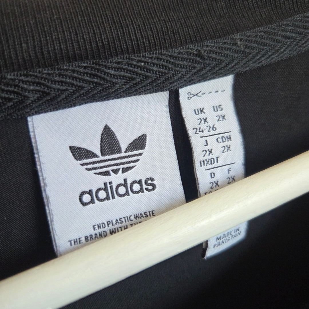 Close-up of two tags inside an XXL Adidas Upcycled Breastfeeding & Pumping Black/White Logo t-shirt (UK24/26). The left tag has the Adidas logo and “End Plastic Waste;” the right shows size info, “Made in Pakistan.” White hanger partly visible.