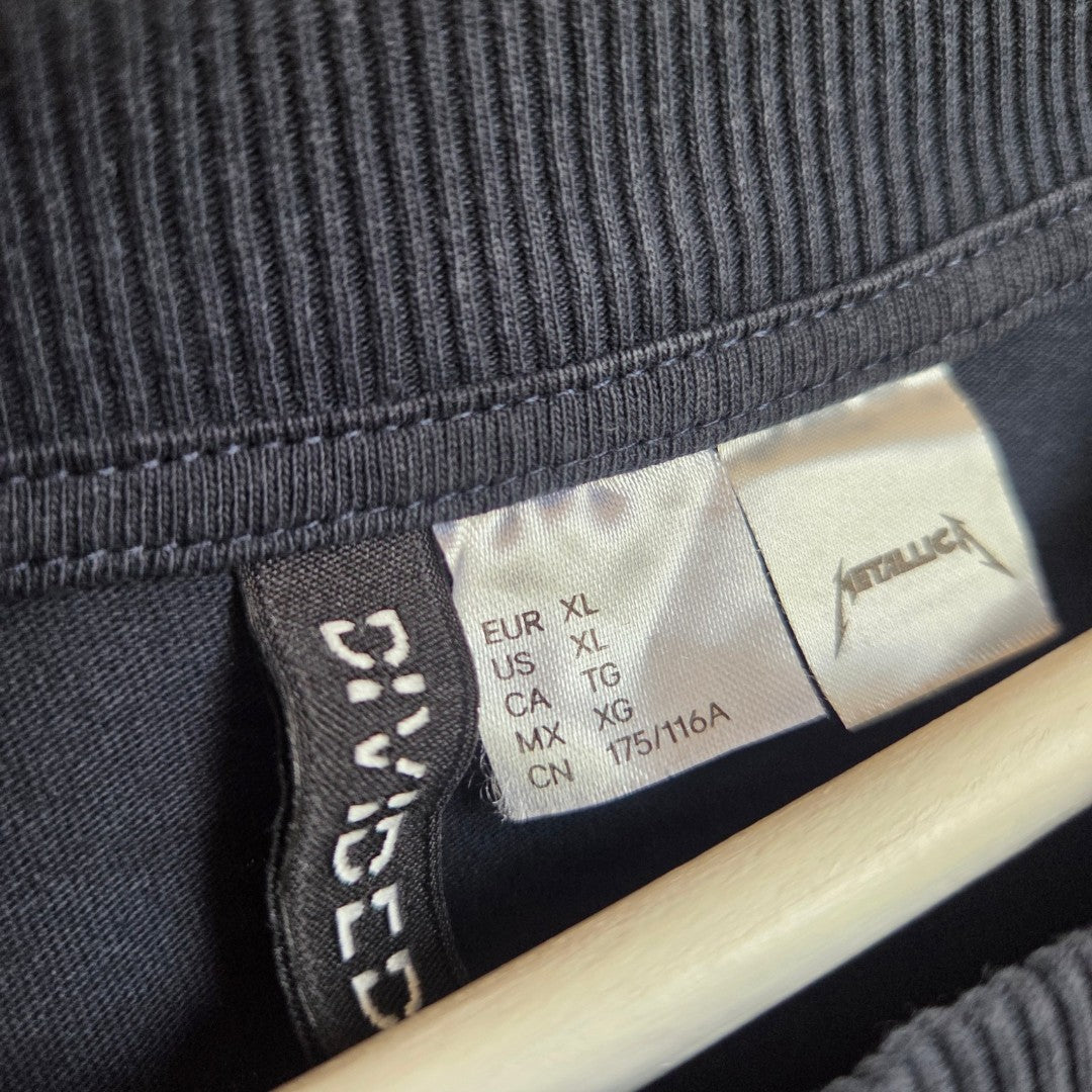 Close-up of an H&M clothing tag on an upcycled navy/green Metallica band t-shirt (XL), featuring size info for multiple regions—a unique, eco-friendly, and affordable nursing & pumping option (100489).