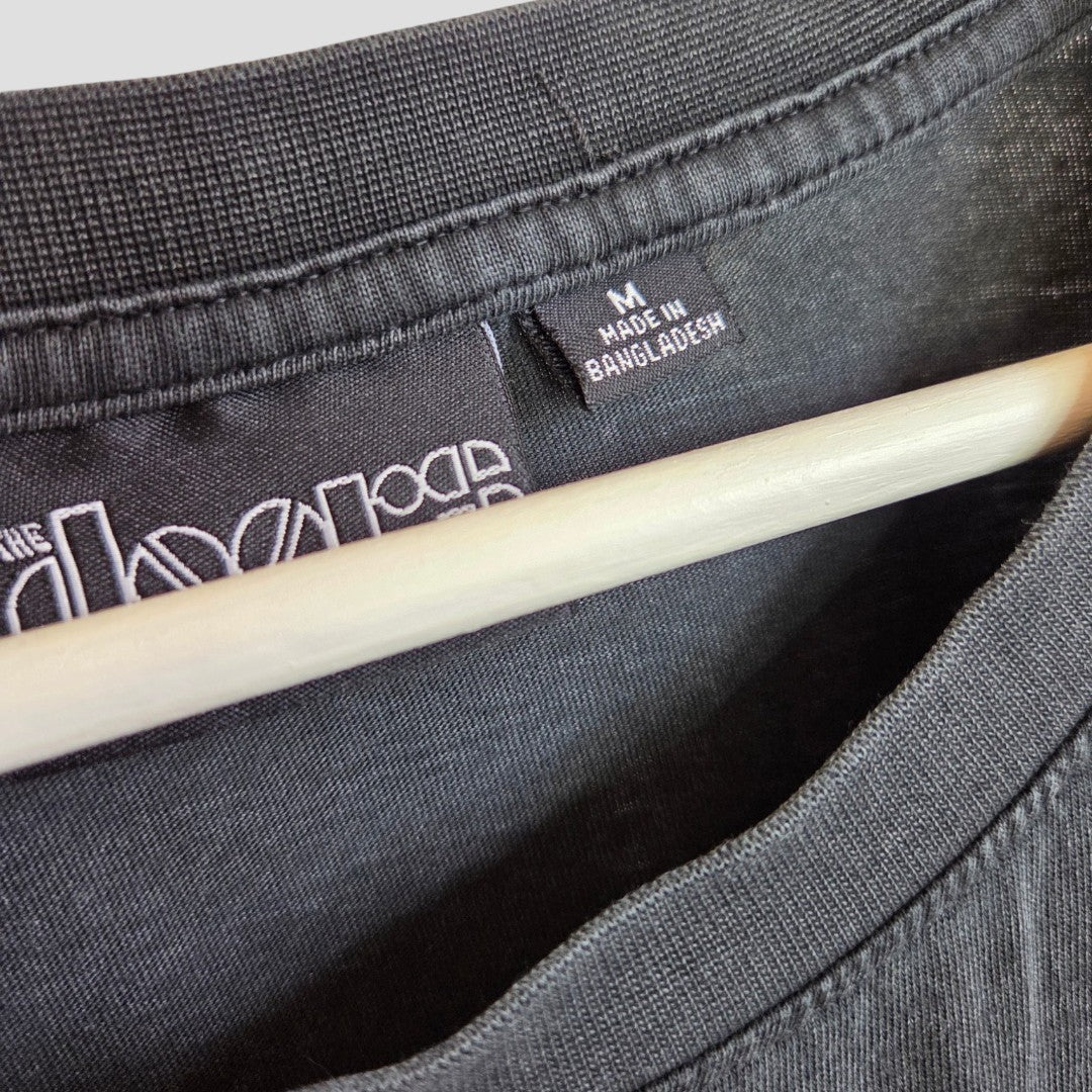 Close-up of the Primark Upcycled Breastfeeding & Pumping The Doors Strange Days Oversized Band t-shirt (M, 100444), black, hanging on a wooden hanger with visible collar, tag labeled M MADE IN BANGLADESH, and partial white print.