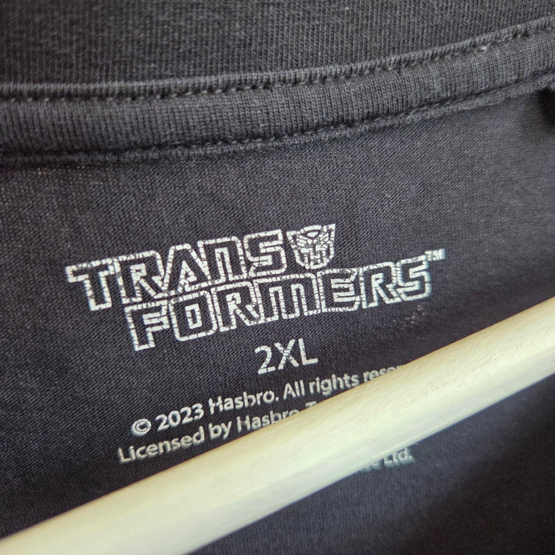 Close-up of the inner label on a black George Upcycled Breastfeeding & Pumping Transformers t-shirt, size 2XL, showing the Transformers logo and Hasbro details. The eco-friendly tee hangs on a hanger, partially visible at the bottom.