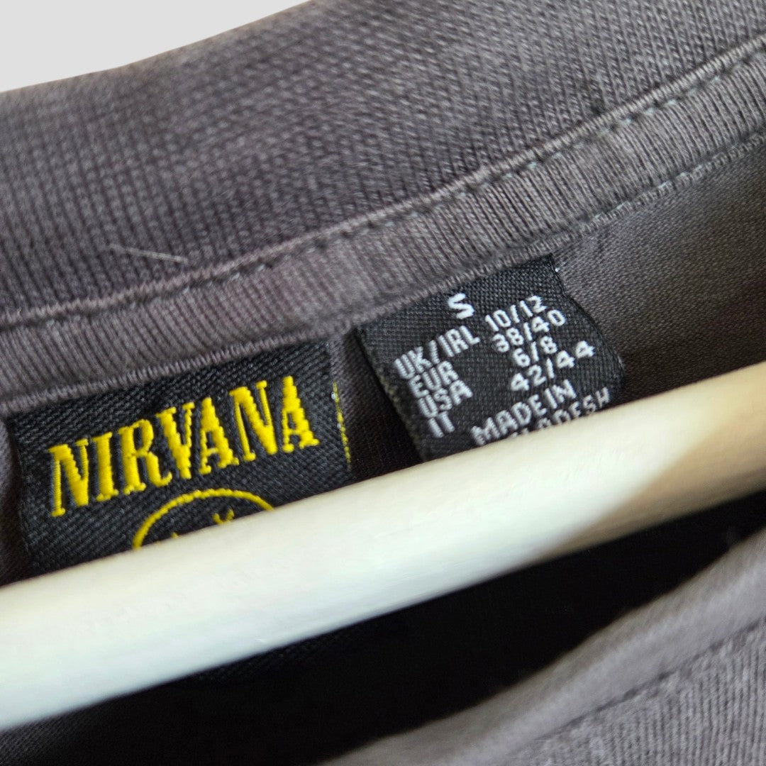 Close-up of a gray Primark Nirvana Yellow Smile Band T-shirt collar (size S/UK10/12), showing two tags—one with yellow Nirvana text, another with size, care info for multiple regions, and MADE IN BANGLADESH—ideal for any nursing wardrobe.