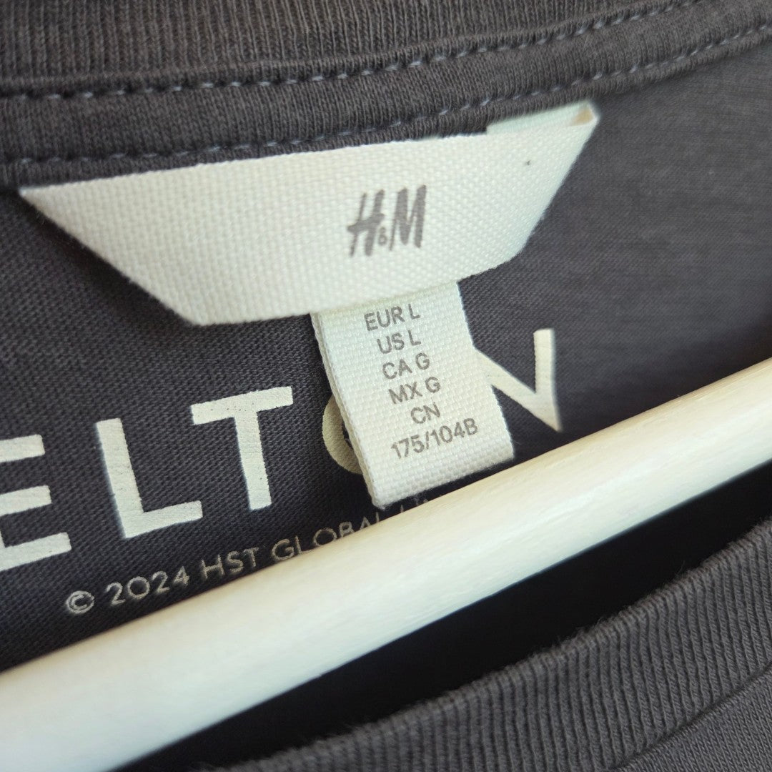 Close-up of an H&M label on the Upcycled Breastfeeding & Pumping Grey Elton John Brick Road Band t-shirt XL, showing sizes: EUR L, US L, CA G, MX G, CN 175/104B. The eco-friendly shirt hangs on a white hanger.