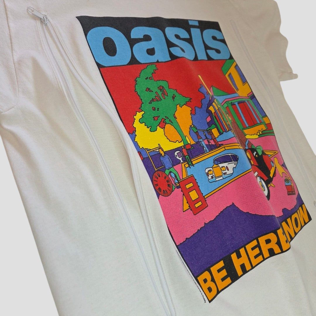 The Gildan Upcycled Breastfeeding & Pumping White Oasis Band t-shirt (M) features a colorful, abstract design with "oasis" at the top and "BE HERE NOW" below. Made from eco-friendly materials for stylish, sustainable comfort.