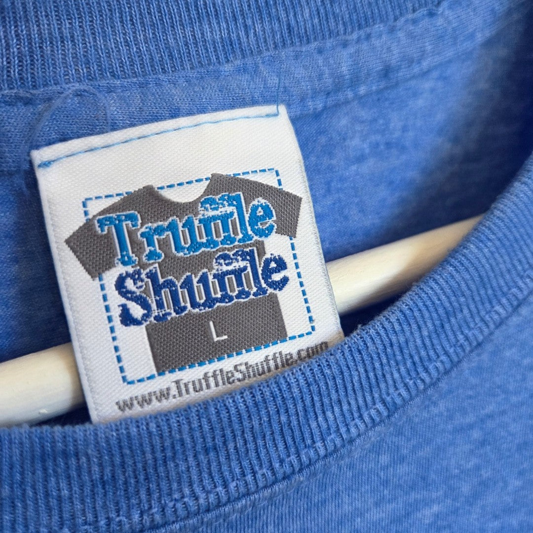 A close-up of a blue shirt’s collar displays a label featuring a T-shirt graphic and the text “Truffle Shuffle L” with www.TruffleShuffle.com printed beneath, highlighting this Upcycled Breastfeeding (Nursing) & Pumping Charlie and the Chocolate Factory t-shirt by Truffle Shuffle—an eco-friendly, affordable piece designed to fit seamlessly into any modern wardrobe.