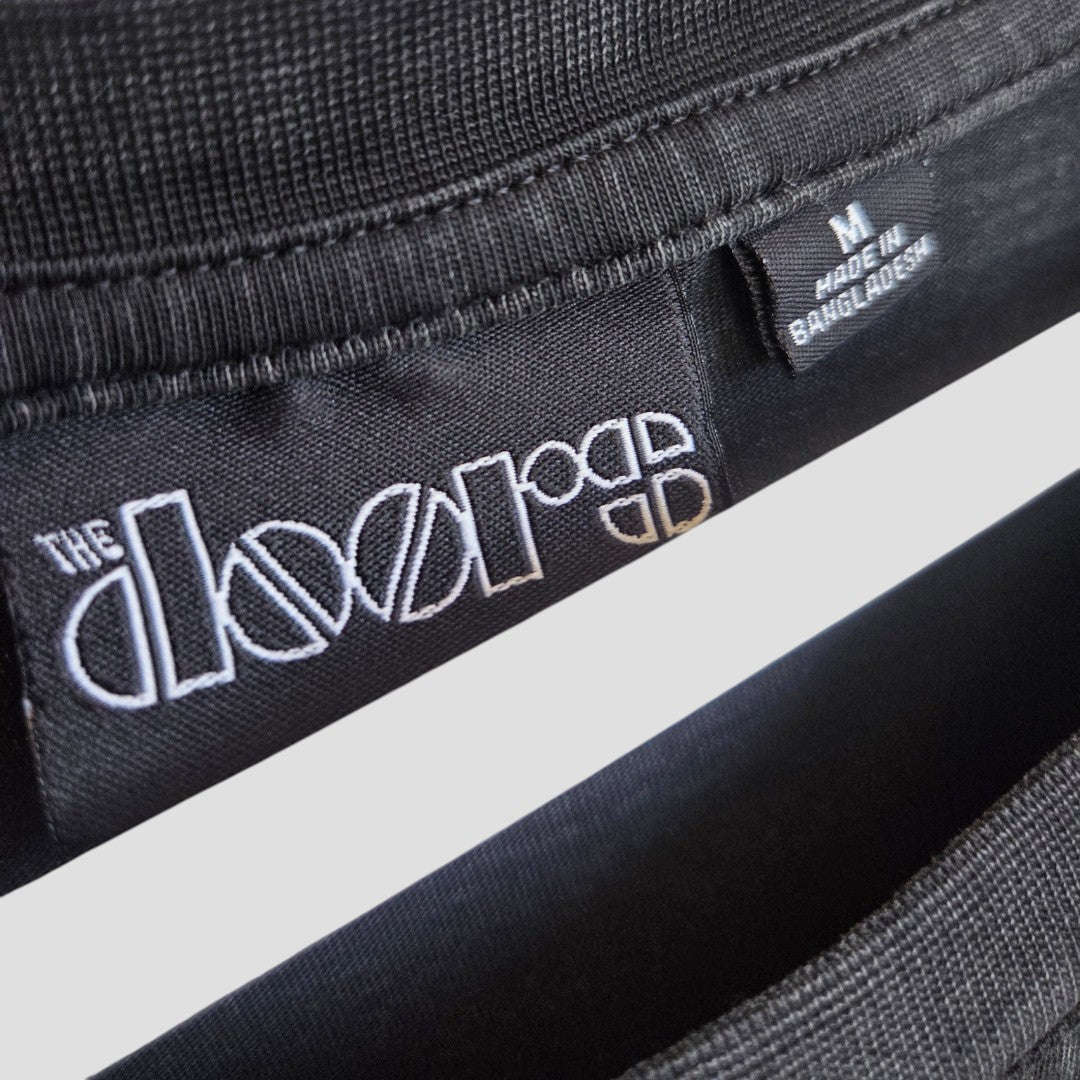 Close-up of black fabric clothing tags on an upcycled Primark knit t-shirt. One tag features The Doors logo, while the other displays "M" and "Made in Bangladesh" in white text. The material appears soft and cozy, making this affordable Upcycled Breastfeeding (Nursing) & Pumping Black/Pink The Doors Band t-shirt (size M, style 100510) a great option for comfortable nursing wear.