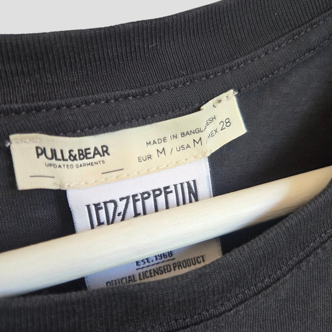 Close-up of the neckline of a black Pull&Bear Upcycled Breastfeeding (Nursing) & Pumping Led Zeppelin Wembley Band t-shirt (size M, 100575), displayed on a hanger. The garment features two tags: one with the Pull&Bear brand and sizing details, and another certifying it as an official licensed Led Zeppelin product. This unique piece combines stylish comfort with affordable, eco-conscious design for everyday wear.