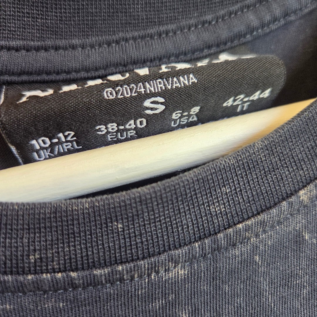 Close-up of a black clothing tag inside a grey/turquoise Nirvana Angel Band nursing & pumping t-shirt by Primark, size S (UK 10/12), showing ©2024 NIRVANA and size equivalents for UK/IRL, EUR, USA, and IT. This eco-affordable upcycled shirt is hanging on a white hanger.