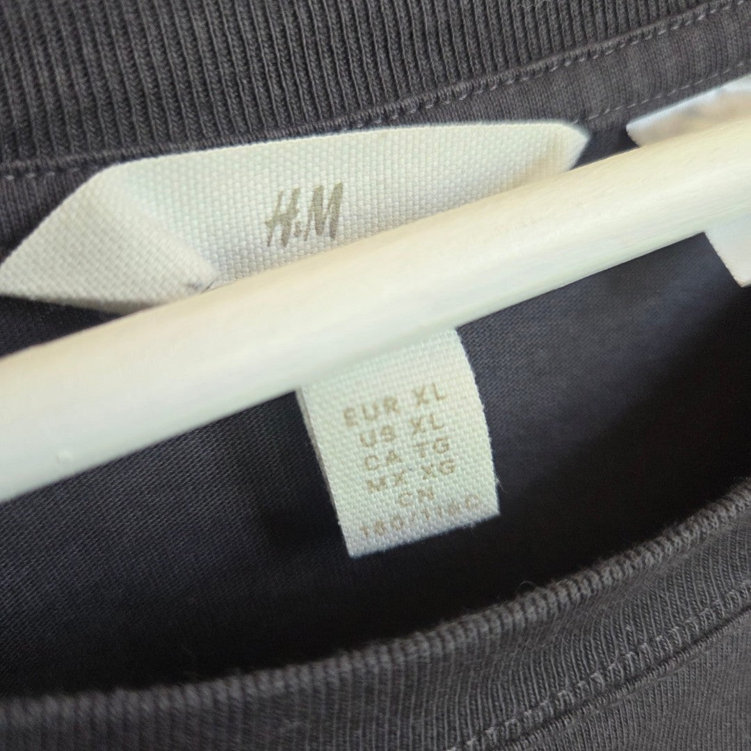 Close-up of an eco affordable Upcycled Breastfeeding (Nursing) & Pumping Grey/Cream Nirvana Snake Band t-shirt XL by H&M on a white hanger, focusing on the inner size tag showing EUR XL, US XL, CA XL, CN 185/112A, and MX XG.