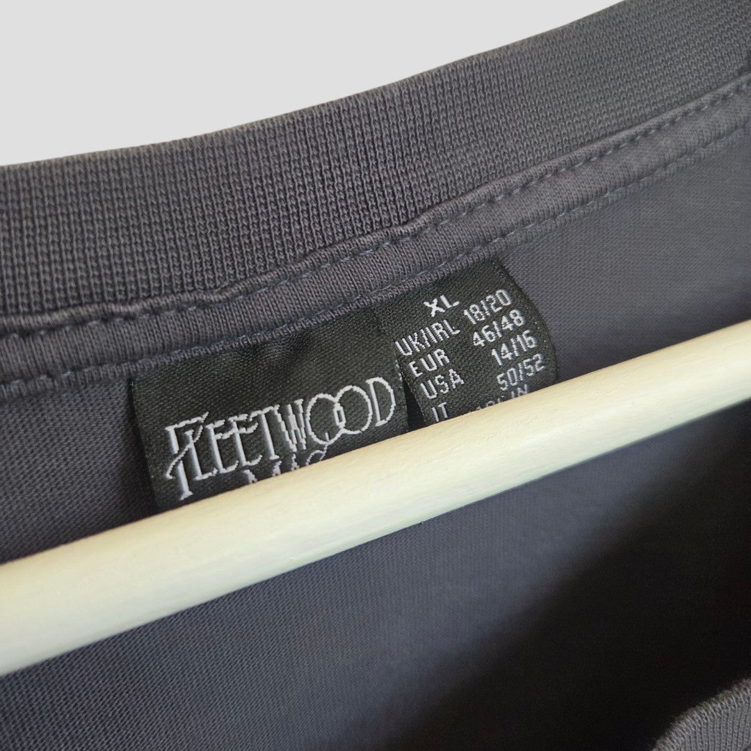 Close-up of a black clothing tag on a dark shirt, reading “Fleetwood” and listing sizes: XL, UK/IRL 18/20, EUR 46/48, USA 14/16, IT 50/52. The shirt—a Primark Upcycled Breastfeeding & Pumping Floral Fleetwood Mac Band t-shirt (XL / UK18/20)—hangs on a light-colored wooden hanger as an affordable nursing clothing option.