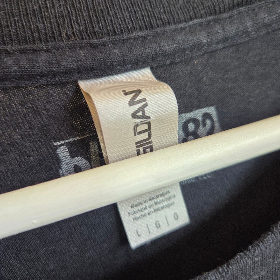 Close-up of the collar on a black Gildan shirt reveals two white tags—one featuring the Gildan brand name and the other listing manufacturing details and size L/G/G, made in Nicaragua. This Upcycled Breastfeeding (Nursing) & Pumping Black Blink-182 Stereo Band t-shirt (L, 100611) is an excellent choice for those seeking affordable nursing wear that fuses daily comfort with unique style.