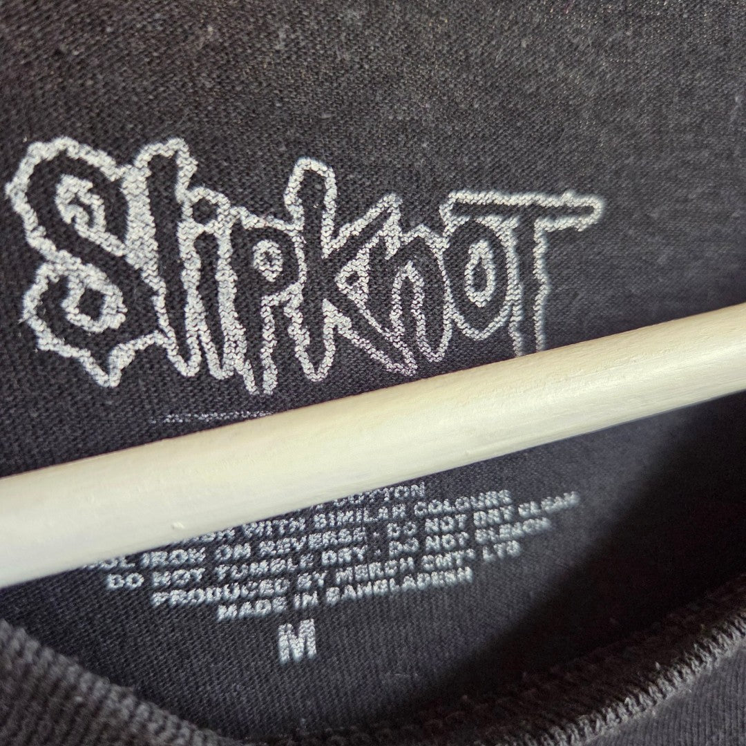 Close-up of the inner label on an upcycled Slipknot band t-shirt (size M) in black/blue, featuring the iconic logo and washing instructions; a wooden hanger is partially visible—offering an edgy, affordable option for unique breastfeeding and pumping wear.