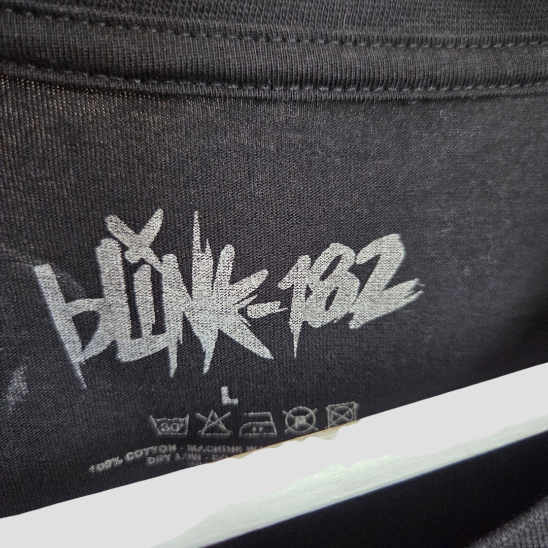Close-up of a black shirt tag from the Blink-182 Upcycled Breastfeeding & Pumping Black/Yellow Smile Band t-shirt (size L), featuring “BLINK-182” in graffiti-style white text above size and care symbols.